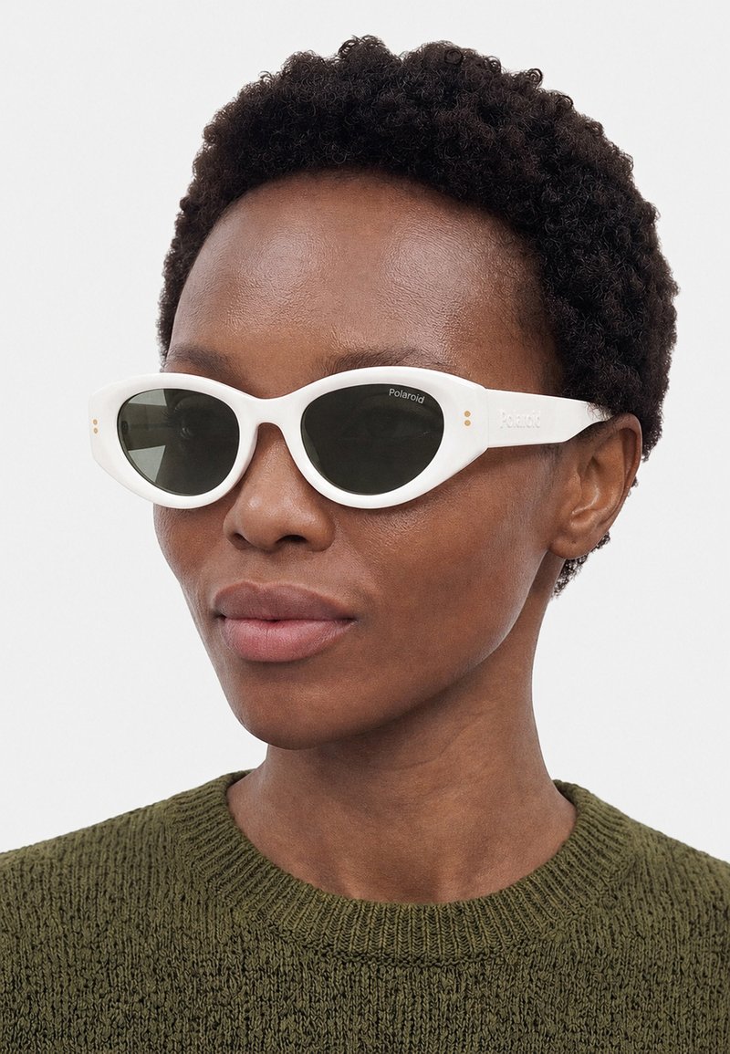 Femme aux cheveux courts et bouclés portant des lunettes de soleil ovales blanches Polaroid et un pull en tricot vert olive, regardant légèrement vers la droite.