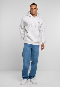 Grauer Hoodie mit Kordeln und einer Vordertasche, kombiniert mit lockeren blauen Jeans und weißen Turnschuhen, vor einem schlichten Hintergrund.