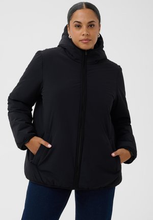 Femme aux cheveux attachés en arrière, portant une doudoune noire à capuche, les mains dans les poches et un jean bleu foncé sur un fond gris uni.