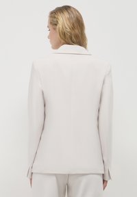 Hellgrauer Blazer mit strukturiertem Kragen, langen Ärmeln und taillierter Passform. Glatte Textur mit seitlichen Belüftungen am Rücken.