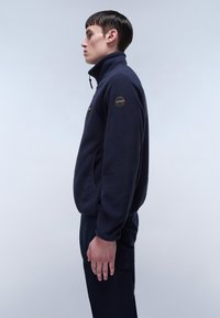 Marineblaues Fleece-Jacke mit hohem Kragen, Seitentaschen und einem runden Emblem am Ärmel. Weiche Textur mit lässiger Passform.