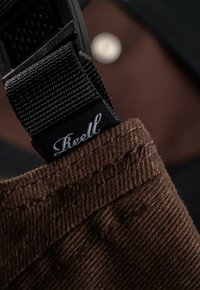 Tela de pana marrón con una textura prominente, que presenta una correa ajustable negra y una etiqueta con un texto en blanco que dice "Reell."