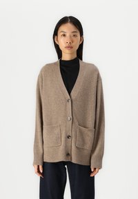 Beige kardigan vyrobený z měkkého materiálu, s výstřihem do V, knoflíkovým zapínáním a dvěma předními kapsami. Rukávy a lem jsou žebrované.