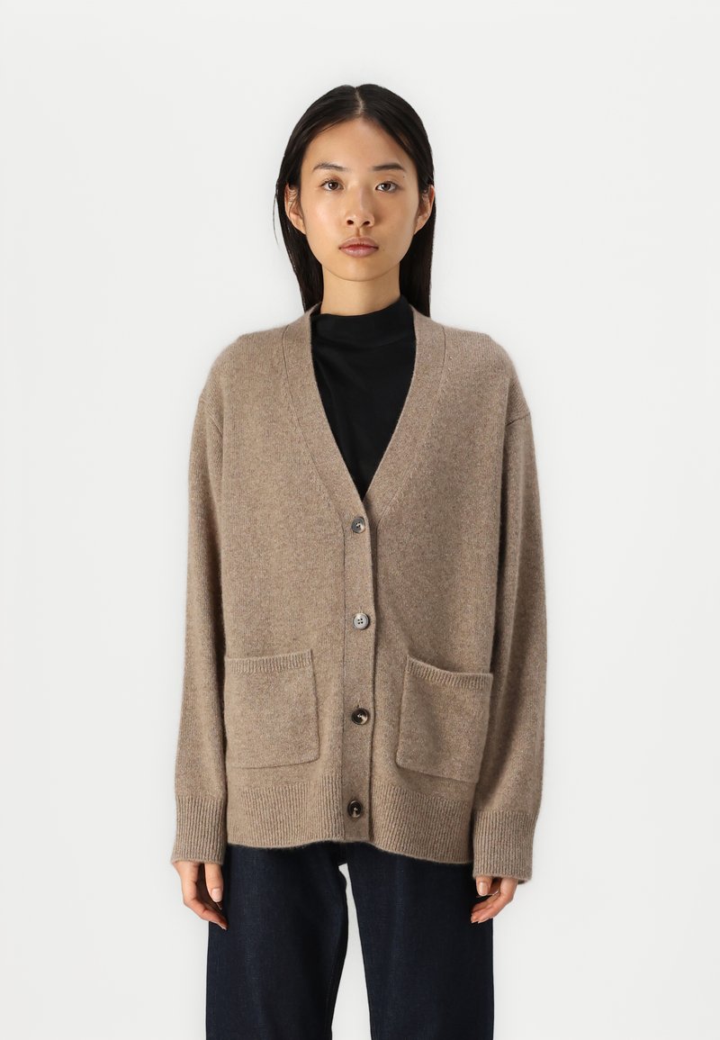 Beige kardigan vyrobený z měkkého materiálu, s výstřihem do V, knoflíkovým zapínáním a dvěma předními kapsami. Rukávy a lem jsou žebrované.