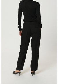 Černé kalhoty na míru s vysokým pasem, rovnými nohavicemi a dvěma zadními kapsami. Nosené s černými podpatkovými mules, z pohledu zezadu.