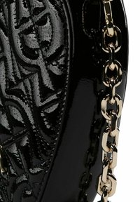 Bolso de mano de charol negro con patrón en relieve, forma curva y asa de cadena dorada con eslabones grandes. Textura lisa y brillante.