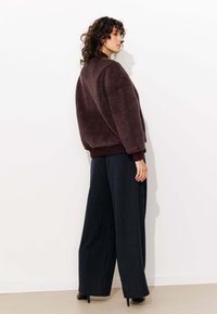 Veste en peluche violette foncée avec des poignets côtelés, portée sur un pantalon large à rayures. Cheveux bouclés et profil latéral visibles sur un fond uni.