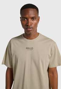 Beige bomullst-shirt med korta ärmar, med svart "HALO"-logotyp tryckt i mitten. Mjuk textur, avslappnad passform.