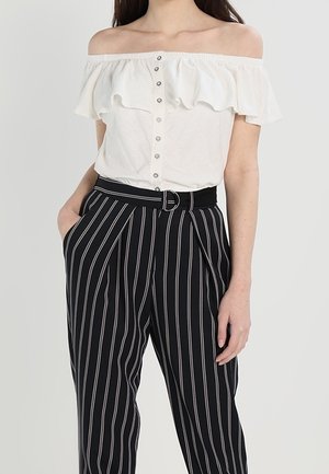 Frau trägt eine schulterfreie weiße Bluse mit Knöpfen und eine hochtaillierte schwarze Hose mit weißen vertikalen Streifen und einem Gürtel.