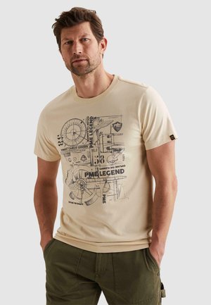 Mann mit kurzem braunem Haar und Bart, trägt ein beiges T-Shirt mit Grafik und olivgrüne Hose, steht mit den Händen in den Taschen vor neutralem Hintergrund.