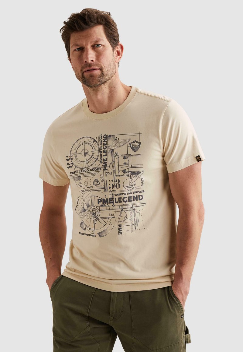 Mann mit kurzem braunem Haar und Bart, trägt ein beiges T-Shirt mit Grafik und olivgrüne Hose, steht mit den Händen in den Taschen vor neutralem Hintergrund.