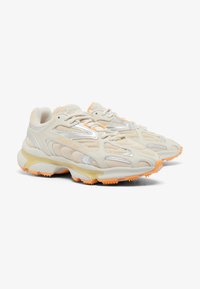 Zapatillas beige con acentos plateados y detalles en naranja. Parte superior de malla con un diseño texturizado, que presenta una suela acolchada y una suela con patrón.