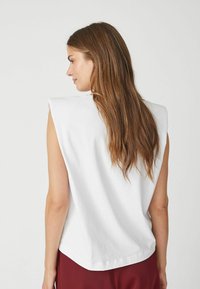 White sleeveless top with a rounded hem and shoulder pads, made of smooth fabric. Long, straight hair falls over the shoulder.