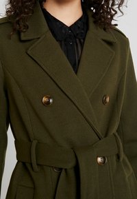 Olijfgroene, dubbelbreasted jas met een riem, gemaakt van gestructureerde stof. Het heeft grote bruine knopen en een ingesneden kraag. Zwarte blouse eronder.
