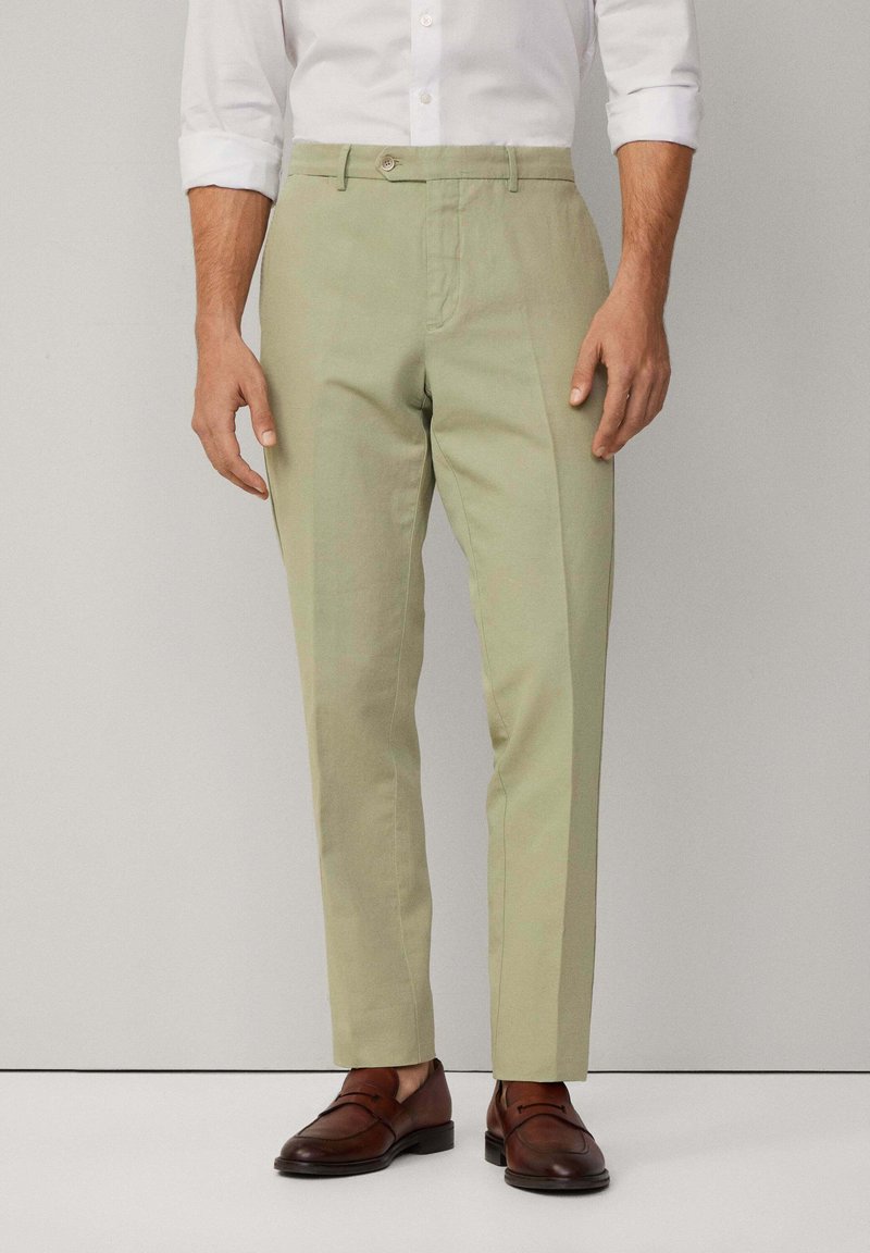 Pantaloni su misura verde chiaro, con una texture liscia, pieghe frontali e chiusura con bottone. Indossati con mocassini in pelle marrone.