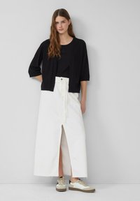 Zwarte cropped cardigan over een zwart top, gecombineerd met een witte maxi rok met een voorkap. Schoenen: beige en witte sneakers.