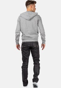 Sweat à capuche gris avec cordon de serrage, associé à un jean noir ajusté avec poches zippées et surpiqûres contrastantes. Baskets blanches visibles.