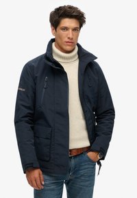 Marineblauwe waterdichte jas met twee met rits afgesloten voorkenhullen, hoge kraag en verstelbare manchetten. Draagt over een crèmekleurige coltrui.