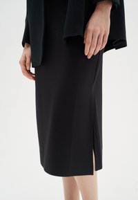 Musta istuva mekko, jossa on sivuveto, valmistettu sileästä kankaasta. Sen helma ulottuu polveen ja siinä on minimalistinen muotoilu.