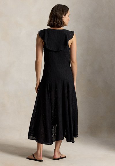 Naine kannab musta varrukateta midi-kleit silmkoelise kangaga ja volangiliste detailidega, seisab paljajalu neutraalse tausta ees.