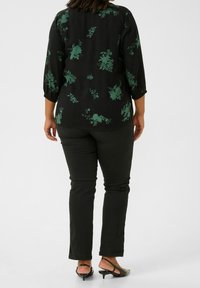 Sort bluse med grønt blomstermønster, trekvartærmer og rund halsudskæring, parret med sorte bukser og lavhælede sko.