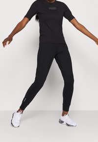 Schwarzes kurzärmeliges T-Shirt mit dezentem Logo, kombiniert mit schwarzen, zulaufenden Jogginghosen mit Seitennähten, getragen mit weißen Sneakers mit gemusterten Sohlen.