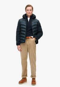Giubbotto imbottito blu scuro con cappuccio, indossato sopra un maglione blu navy. Pantaloni di corduroy beige e stivali marroni con lacci gialli completano l'outfit.
