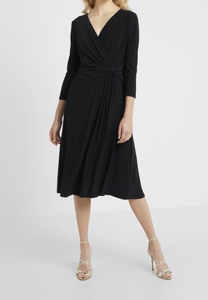 Femme portant une robe midi noire style portefeuille, manches trois-quarts, et des sandales à talons hauts dorées à brides.