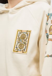 Κρεμ φούτερ με κουκούλα με κεντητό σχέδιο δύο κίτρινων ηλιακών προσώπων και ανθικά μοτίβα στο στήθος, με παρόμοια σχέδια στο μανίκι.