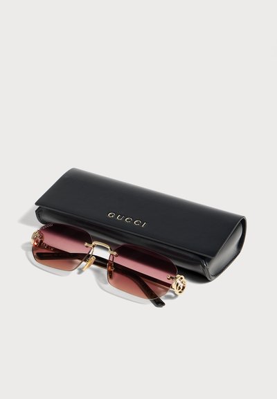 Слънчеви очила Gucci със златиста рамка и розови стъкла са поставени пред черен твърд калъф Gucci на бял фон.