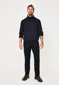 Dunkelblauer Rollkragenpullover mit strukturiertem Strick, kombiniert mit schwarzen Hosen und schwarzen Schnürstiefeln. Das Model steht in einer neutralen Umgebung.