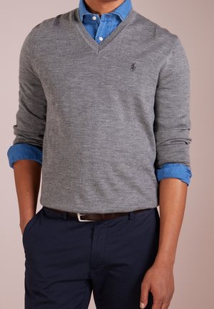 Mann trägt einen grauen V-Ausschnittpullover über einem blauen Hemd mit hochgekrempelten Ärmeln, kombiniert mit dunkelblauer Hose und braunem Gürtel.