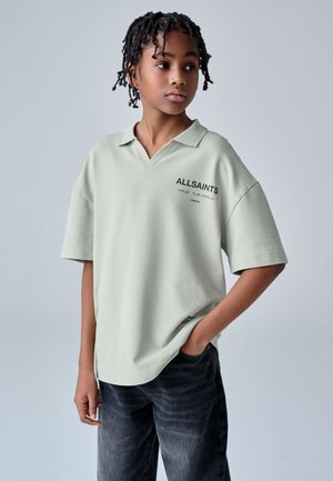 Mladá osoba s copánky, která má na sobě světle šedé oversize polo tričko s nápisem „ALLSAINTS“, stojí s jednou rukou v kapse a dívá se doprava.