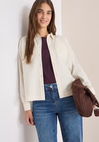 Camicia button-up color crema con due tasche sul petto, indossata sopra un top viola scuro. Abbinata a jeans blu e che tiene in mano una borsa marrone.