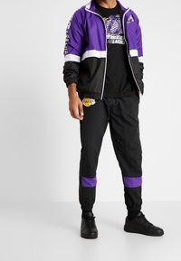 Lila och svart vindjacka med vita ränder och logotyp, kombinerat med svarta byxor med lila detaljer och Lakers-logotyp, bärs med svarta skor.