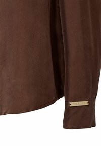 Cămașă din suede maro, cu o textură netedă, margine rotunjită și etichetă din metal pe manșetă gravată cu "PIESZAK."