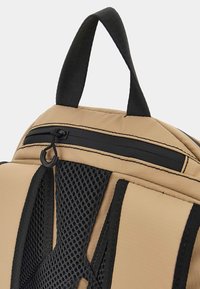 Beige tygryggsäck med svarta nät-accenter. Har ett handtag upptill, svart dragkedja med dragrem och vadderade axelremmar.