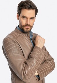 Giacca di pelle marrone con spalle trapuntate, chiusura a pressione e superficie leggermente texturizzata, indossata sopra una camicia blu.