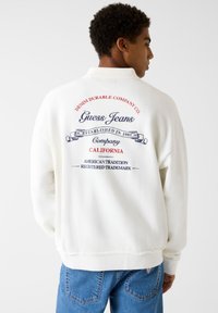 White sweatshirt with front collar, featuring blue and red branded text on the back. Fabric is soft; design includes established date and logo.