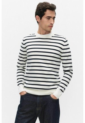 Homme portant une chemise blanche à manches longues avec des rayures horizontales noires et un jean bleu foncé, les mains dans les poches, regardant sur le côté.