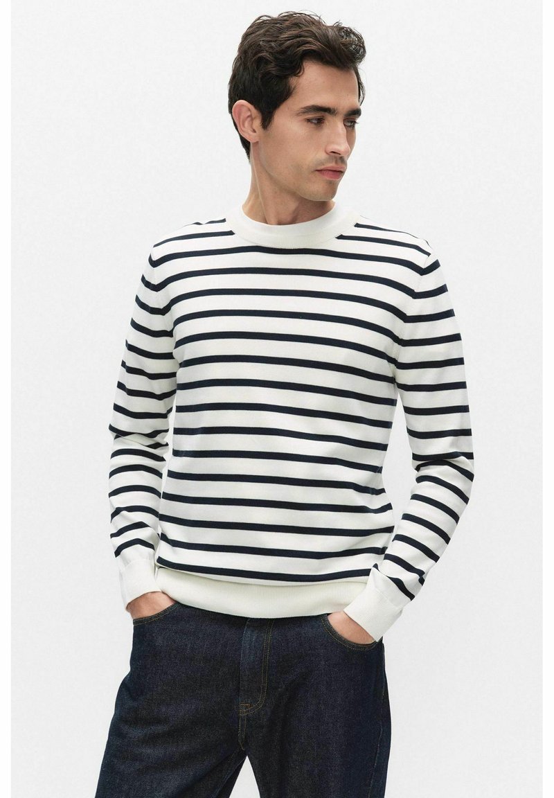 Man met een wit shirt met lange mouwen en zwarte horizontale strepen, en donkerblauwe spijkerbroek, handen in de zakken, kijkt opzij.