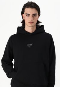 Svart hettegenser laget av mykt stoff med en frontlomme, med en hvit tekstlogo i midten. Designet inkluderer en snor til hetten.