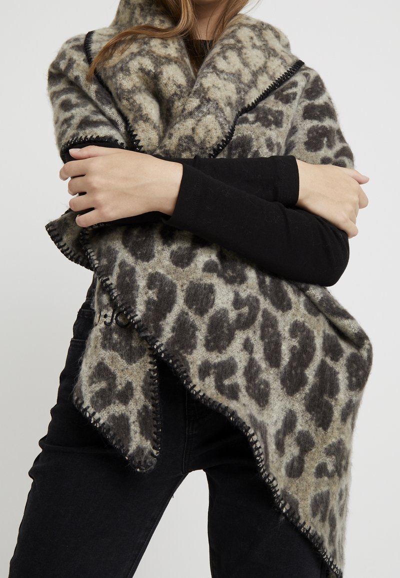 Personne portant des manches longues noires et un pantalon noir, enveloppée dans un grand châle en laine gris et noir à imprimé léopard avec des bords cousus.