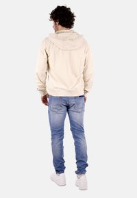 Felpa beige con cappuccio, dalla vestibilità rilassata, con polsini e orlo a costine, abbinata a jeans azzurri chiari e sneakers bianche. Design semplice, senza loghi visibili.