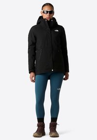 Czarna ocieplana parka z kapturem, dwie zapinane na zamek kieszenie boczne, w połączeniu z turkusowymi legginsami i brązowymi butami turystycznymi. Na nosie okulary przeciwsłoneczne.