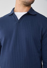 Mornarsko modra, rebrasta polo majica z dolgimi rokavi in V-izrezom ter ovratnikom. Tkanina ima mehak texture, primerna za sproščeno nošenje.