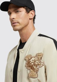 Beige bomberjakke med sorte detaljer, der har et stort broderet tigerpatch, trykknaplukning og ribbet krave.