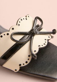 Schwarz-weiße flache Sandale mit einem gewellten Design, die eine große schwarze Schleife und komplexe Perforationen auf dem Obermaterial aufweist.