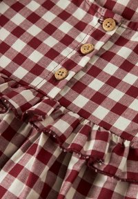 Tela a cuadros en rojo y crema con un dobladillo rizado. Cuatro botones de madera exhibidos de manera prominente a lo largo de la abertura, material texturizado tejido.