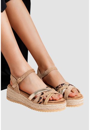 Pieds portant des sandales à plateforme beiges avec semelles en corde tressée, brides métalliques et imprimé léopard, et fermetures à boucle à la cheville sur un fond uni.